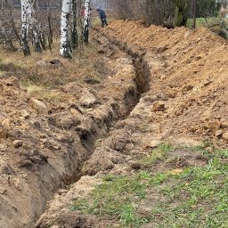 MMIM Budowa Michał Bączyński - Wykopany wąski rów w ziemi, prawdopodobnie pod przyłącza wodociągowe, z widoczną osobą pracującą w oddali i brzozami w tle.
