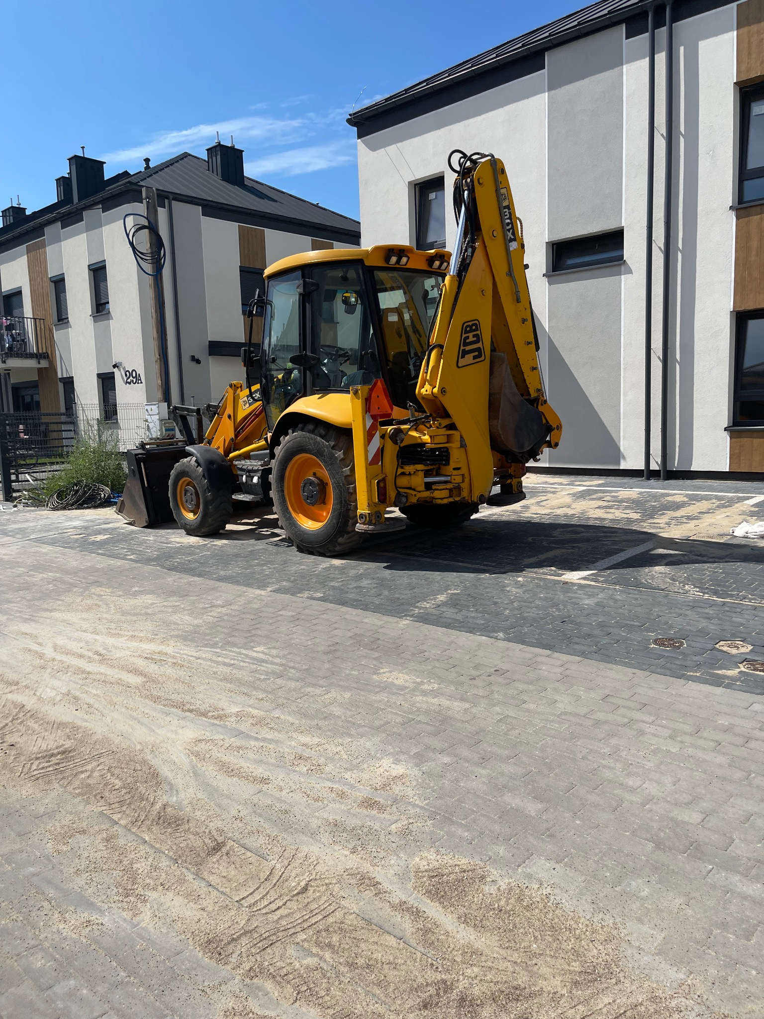 Żółta koparko-ładowarka JCB na brukowanej powierzchni przed nowymi budynkami mieszkalnymi w słoneczny dzień, widoczne ślady opon i piasek na kostce.