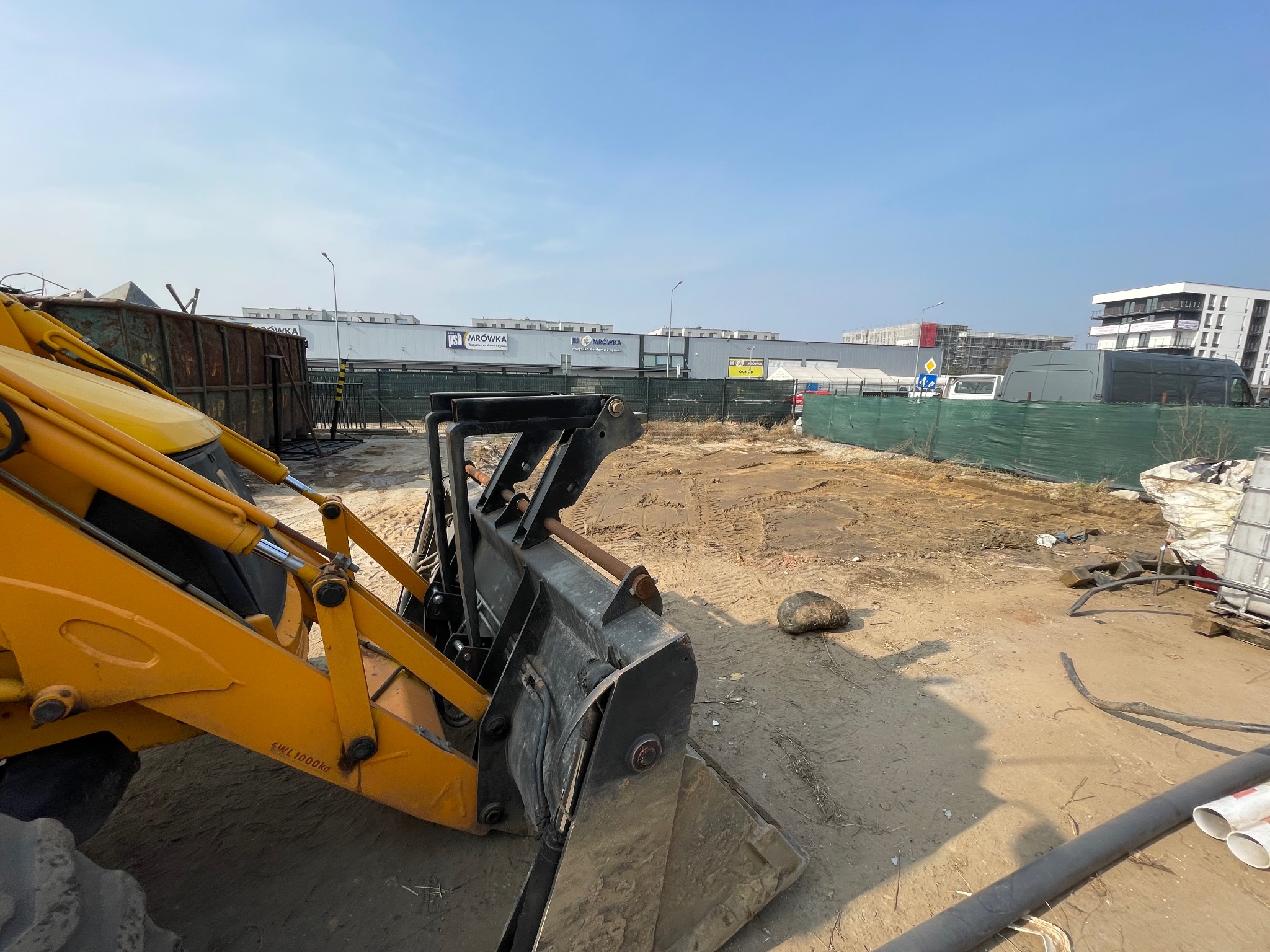 Żółta koparko-ładowarka z czarną łyżką na placu budowy, w tle budynki handlowe i biurowe pod błękitnym niebem.