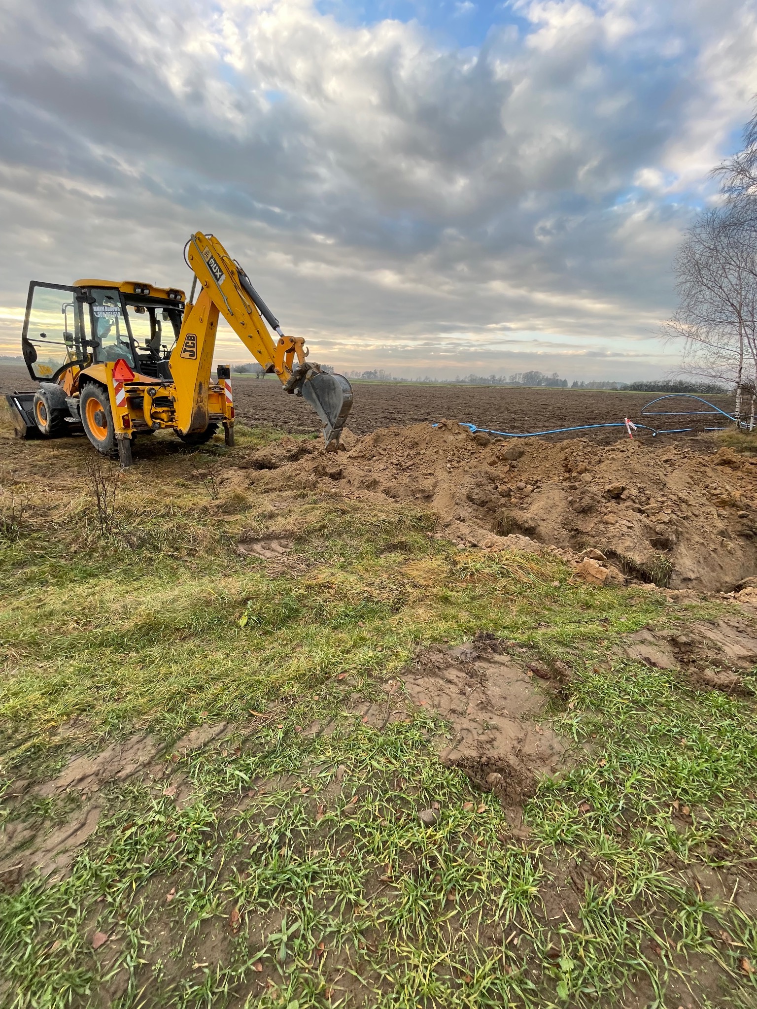 Żółta koparko-ładowarka JCB podczas prac ziemnych na polu, widoczne wykopane rury wodociągowe.