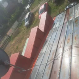 Blaszany dach z widocznymi śladami rdzy, z zamontowaną anteną satelitarną i nową obróbką blacharską komina pomalowaną na czerwono, na tle podwórza z samochodami.