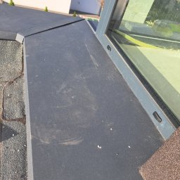 Dach As - Szczegółowe ujęcie płaskiego dachu z szarej papy bitumicznej, przylegającego do okna dachowego z szarą ramą, na tle białego budynku z oknem