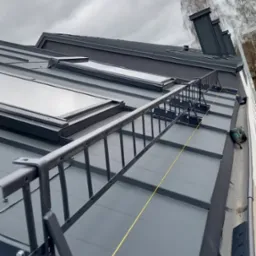 Szary dach z blachy trapezowej z zamontowanymi oknami dachowymi, zabezpieczeniami komunikacyjnymi w postaci drabinki dachowej i linką asekuracyjną, widok z góry na pochmurnym tle.