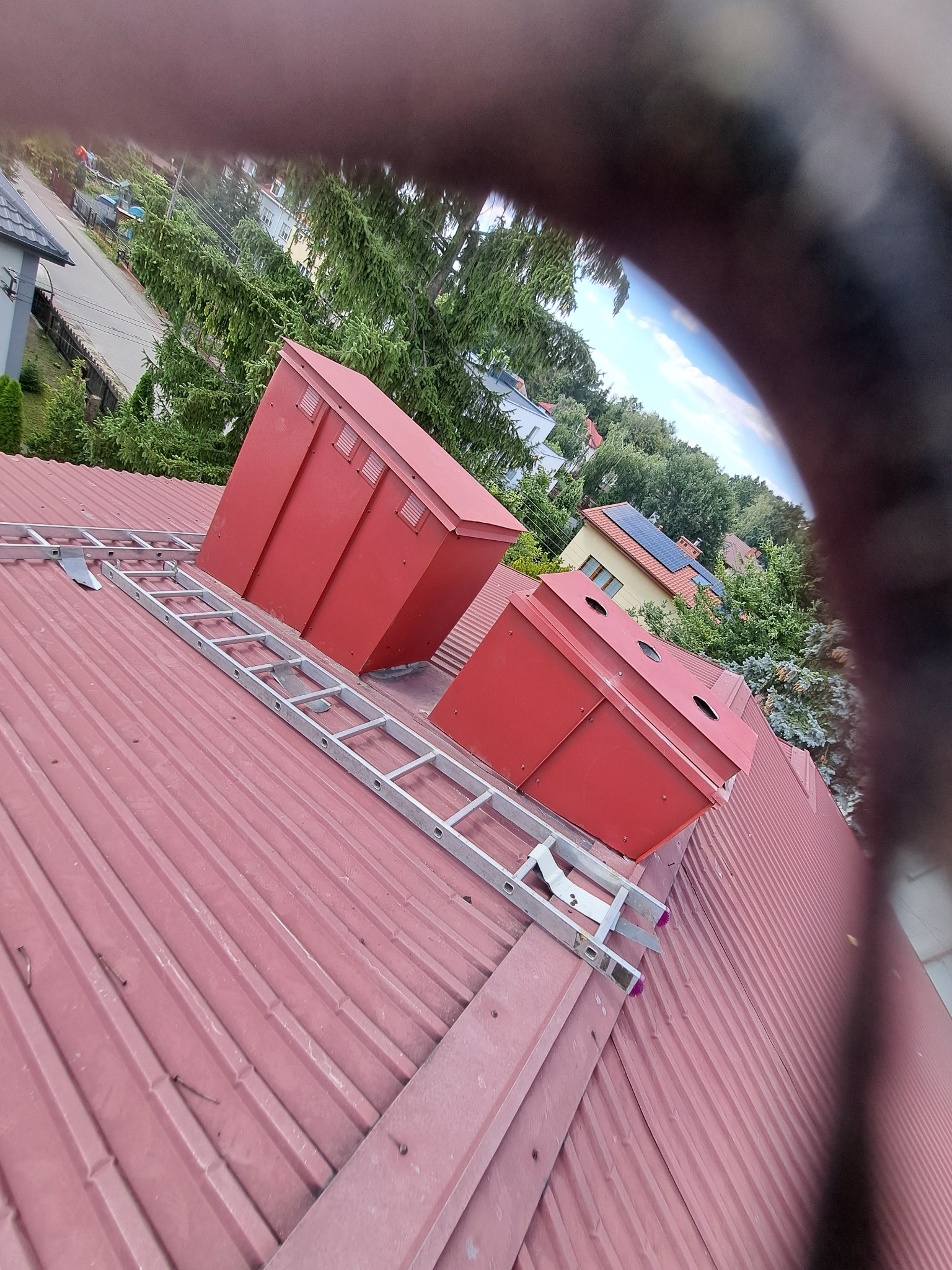 Czerwony dach z blachy trapezowej, na nim drabina dekarska i dwa czerwone kominy wentylacyjne, w tle zabudowa jednorodzinna i zieleń. Zdjęcie zrobione z nietypowej perspektywy z widocznym...