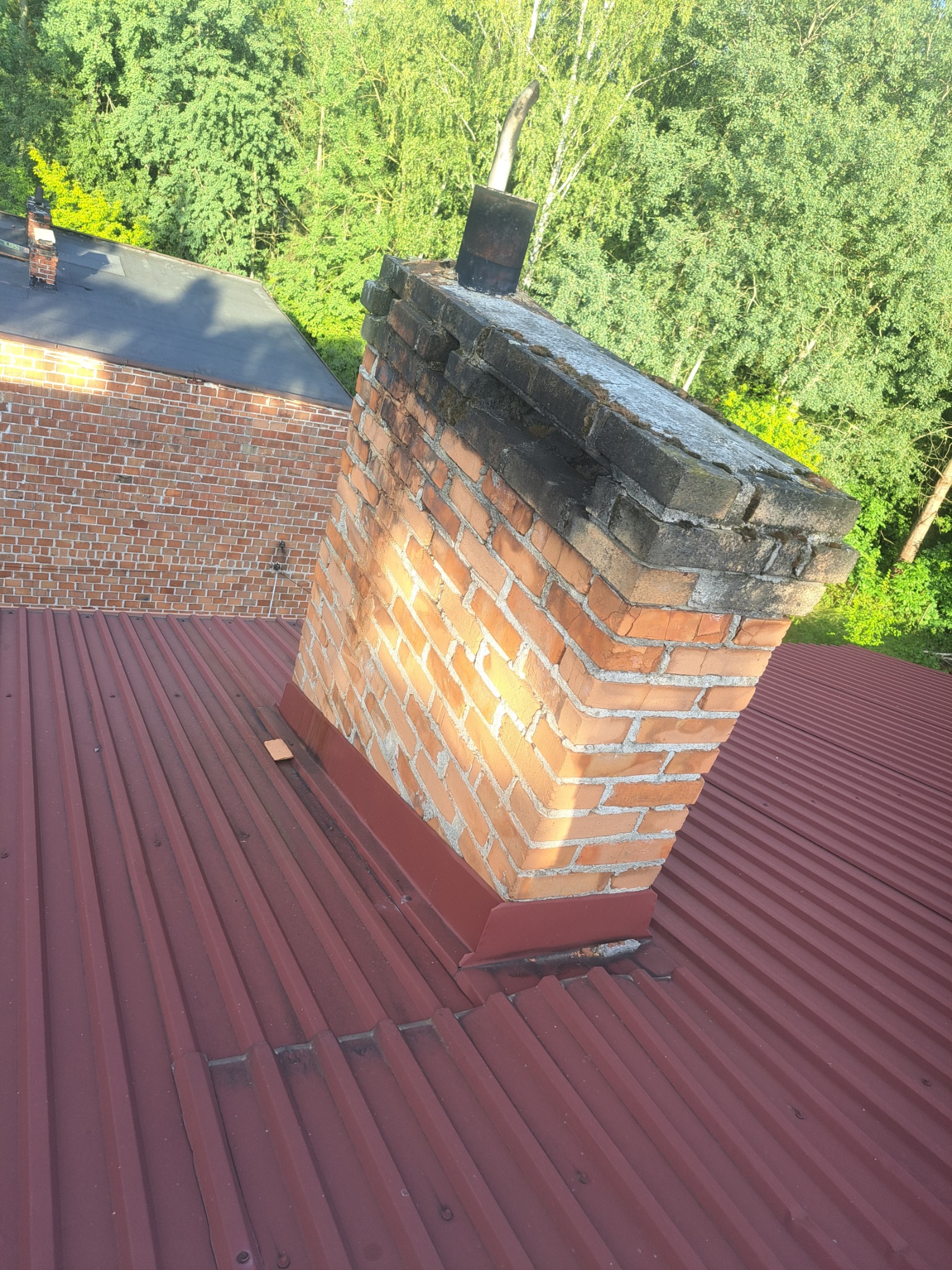 Ceglany komin na czerwonym dachu z blachy falistej, widoczne zabrudzenia i osad na szczycie komina, w tle zielone drzewa.