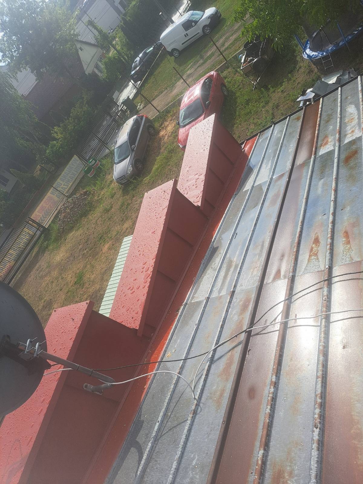 Metalowy dach z widocznymi śladami korozji i świeżo pomalowanymi na czerwono elementami komina, w tle zaparkowane samochody i zieleń.