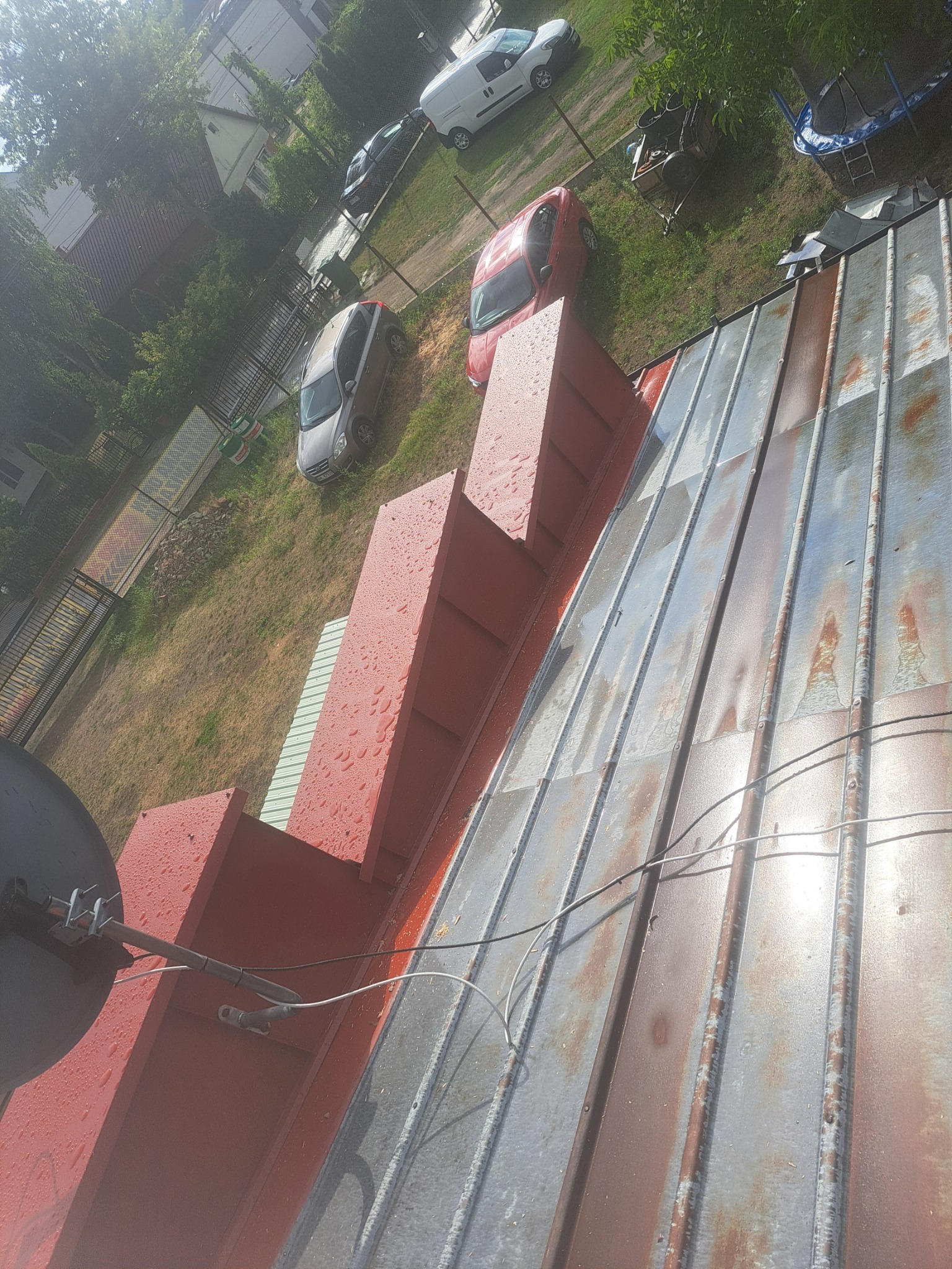 Blaszany dach z widocznymi śladami rdzy, z zamontowaną anteną satelitarną i nową obróbką blacharską komina pomalowaną na czerwono, na tle podwórza z samochodami.