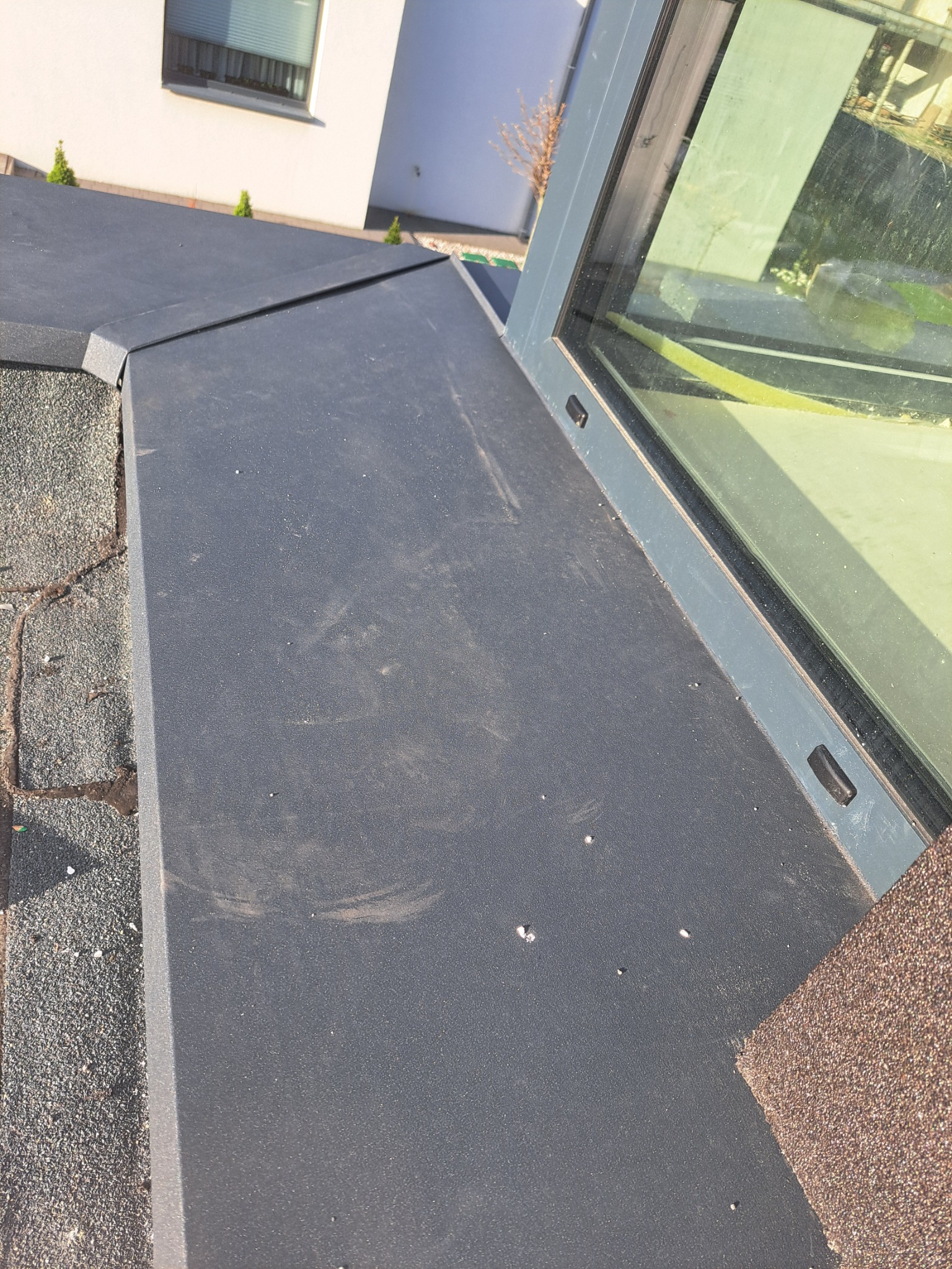 Szczegółowe ujęcie płaskiego dachu z szarej papy bitumicznej, przylegającego do okna dachowego z szarą ramą, na tle białego budynku z oknem