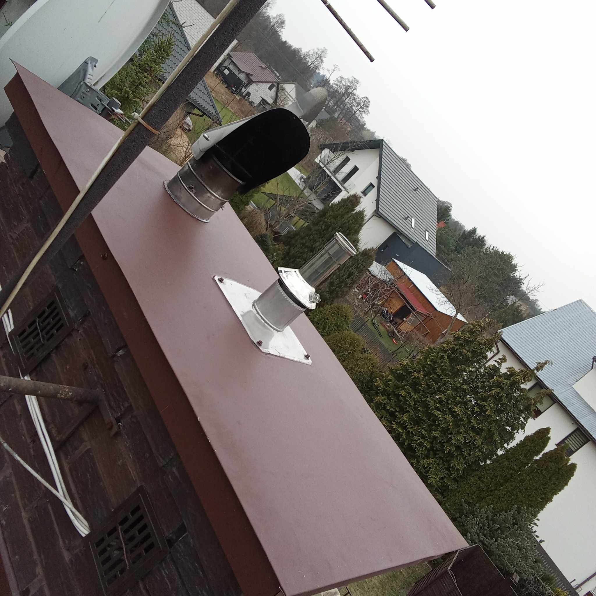 Nowe, brązowe krycie dachu z kominem i odpowietrznikami na tle domów w Otwocku, widok z góry pod kątem.