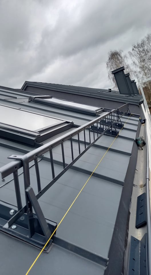 Szary dach z blachy trapezowej z zamontowanymi oknami dachowymi, zabezpieczeniami komunikacyjnymi w postaci drabinki dachowej i linką asekuracyjną, widok z góry na pochmurnym tle.