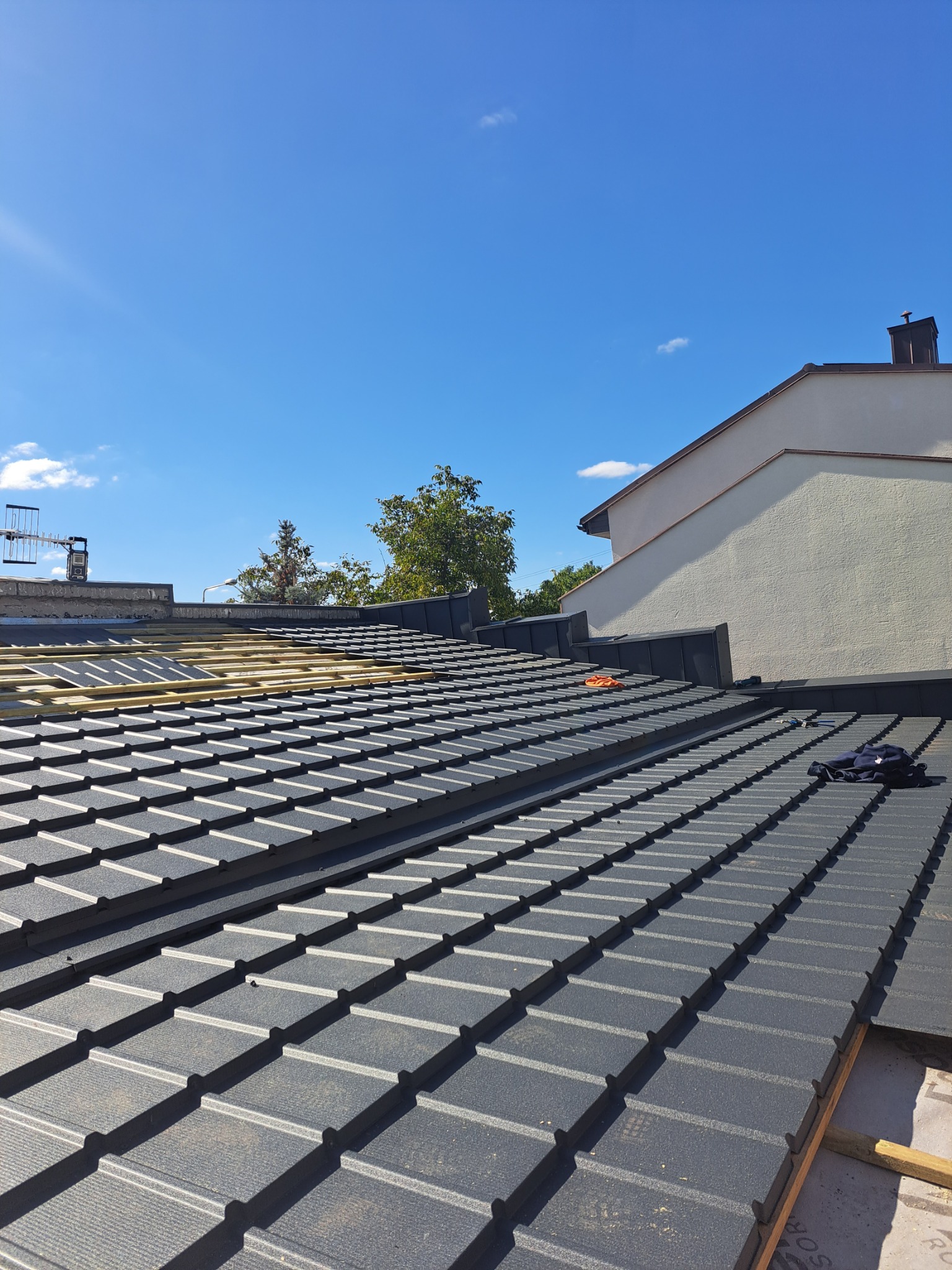 Częściowy widok dachu pokrytego grafitową dachówką modułową, z widocznym fragmentem konstrukcji drewnianej i membraną, na tle błękitnego nieba i fragmentu budynku.