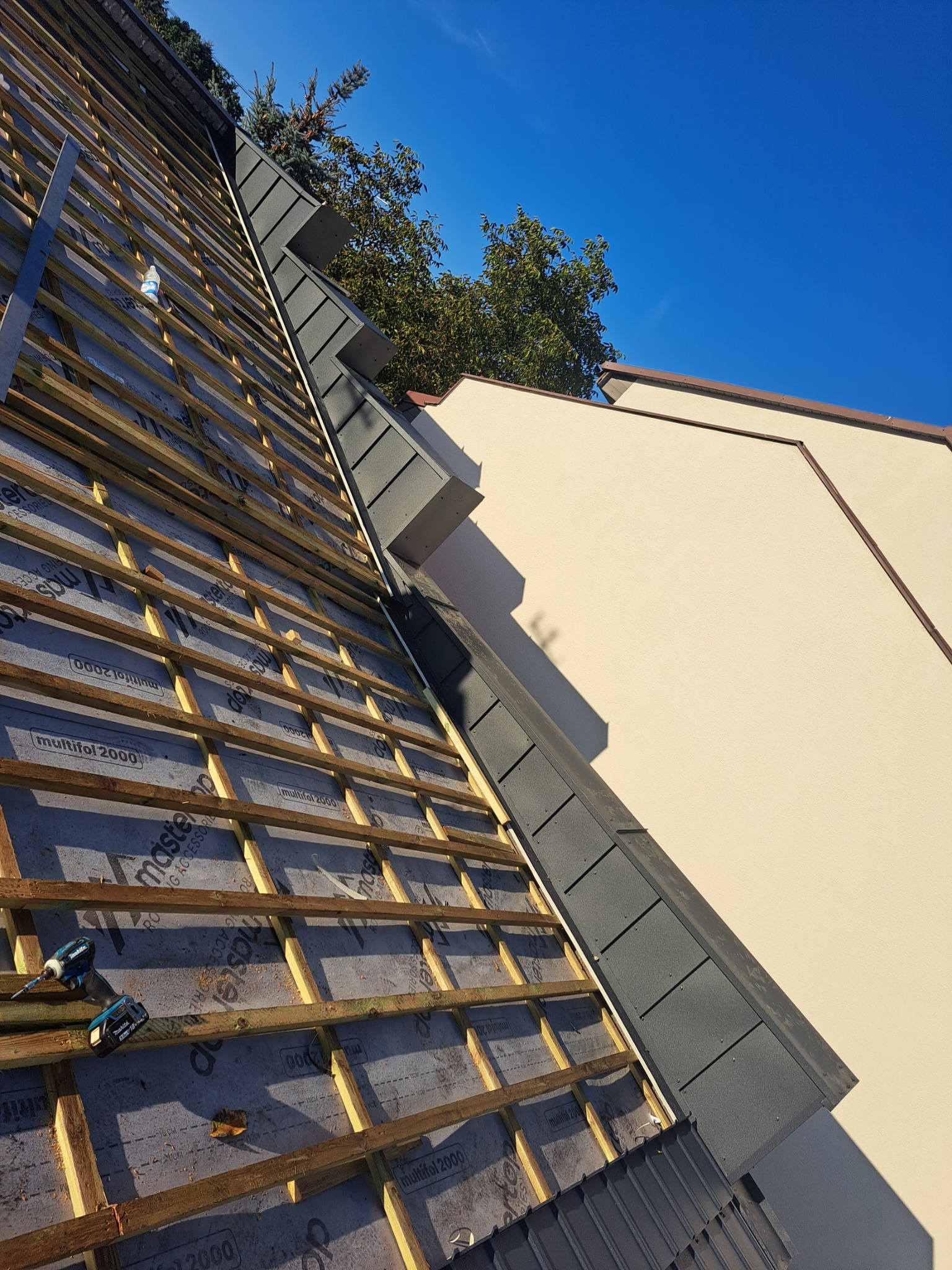 Konstrukcja drewniana pod nowy dach z szarej blachy trapezowej, widoczna membrana dachowa z nadrukiem, w tle fragment elewacji budynku i błękitne niebo.