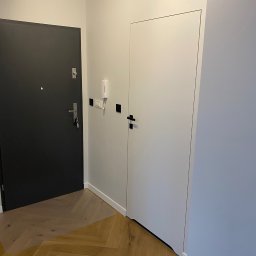 Modern Flat firma remontowa - Korytarz po remoncie z dwiema drzwiami: czarne wejściowe z kluczami w zamku i białe wewnętrzne, parkiet ułożony w jodełkę, ściany pomalowane na biało.