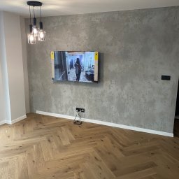 Modern Flat firma remontowa - Salon z parkietem ułożonym w jodełkę, ściana z dekoracyjnym tynkiem, wiszący telewizor i nowoczesna lampa sufitowa z trzema szklanymi kloszami.