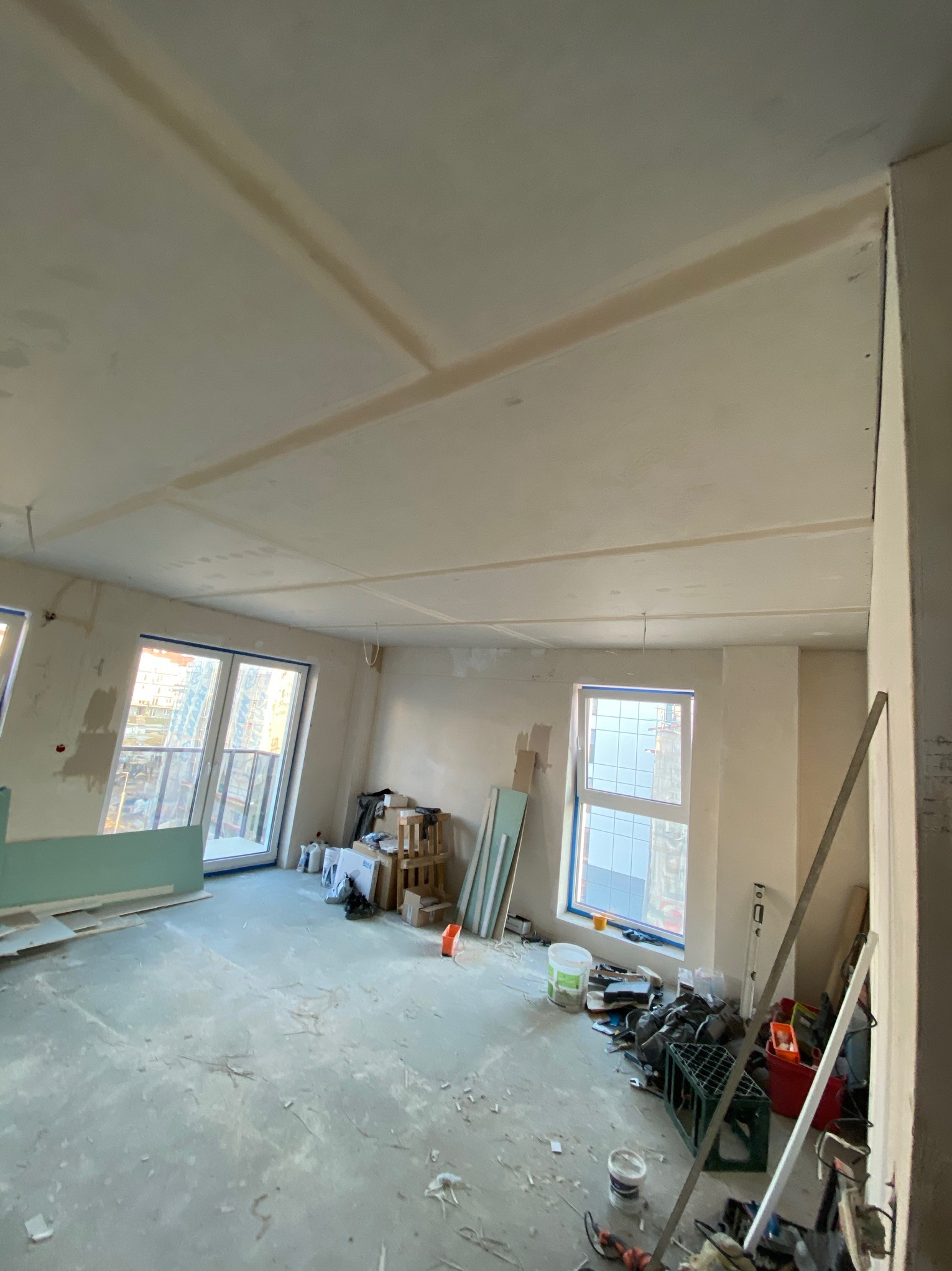 Wnętrze w trakcie prac wykończeniowych: surowy sufit z widocznymi łączeniami płyt kartonowo-gipsowych, przygotowany do dalszej obróbki, podłoga z resztkami materiałów budowlanych, okna z widokiem...