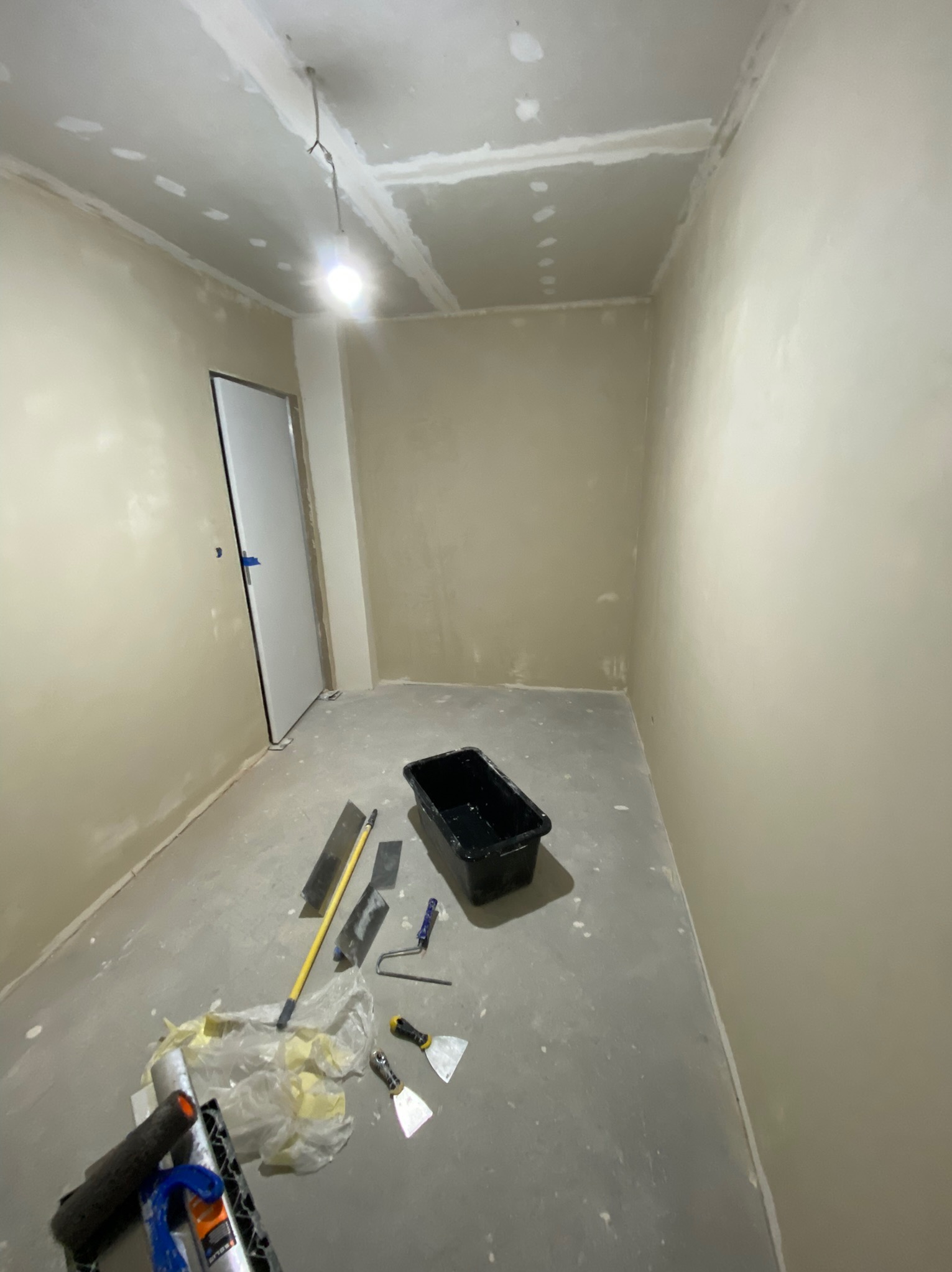 Pokój w trakcie prac przygotowawczych do wykończenia, ściany po szpachlowaniu, widoczne narzędzia malarskie i wiadro na podłodze, surowe oświetlenie.