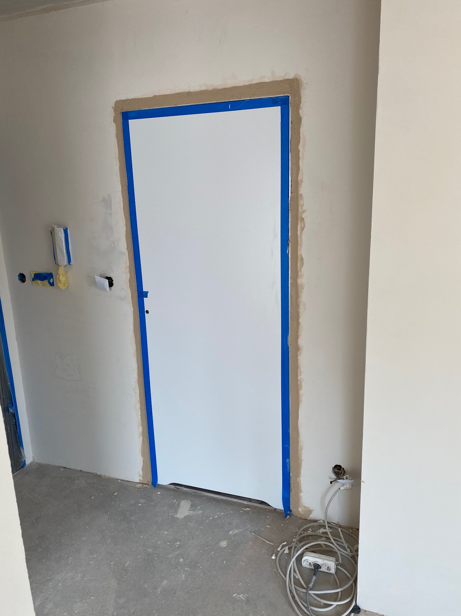 Białe drzwi z niebieską taśmą malarską zabezpieczającą ościeżnicę podczas prac wykończeniowych, widoczne gniazdko elektryczne z przedłużaczem i niedoskonałości na ścianie przed malowaniem.