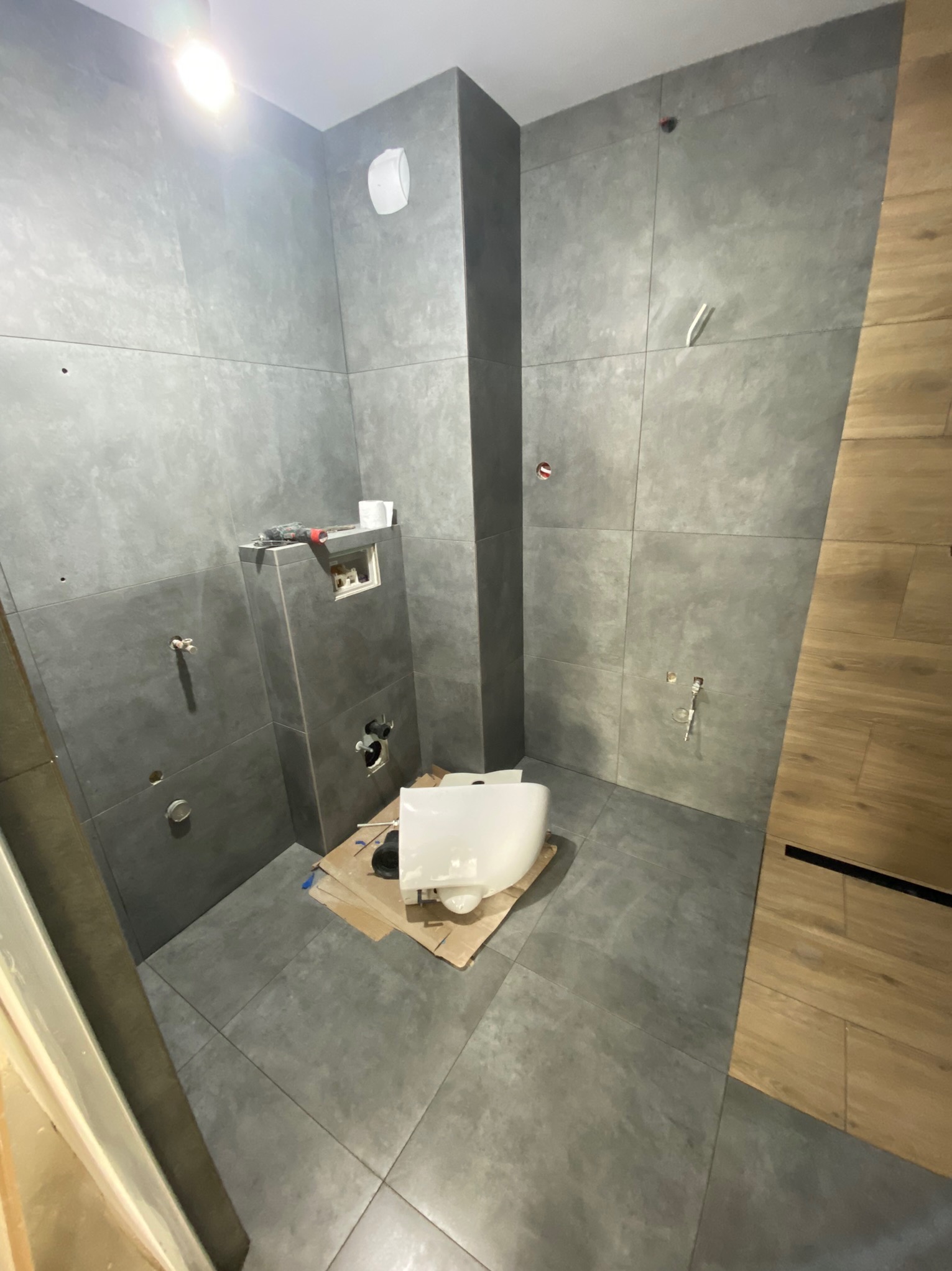 Wnętrze łazienki w trakcie montażu podwieszanej toalety na tle szarych płytek; widoczne narzędzia i instalacje.