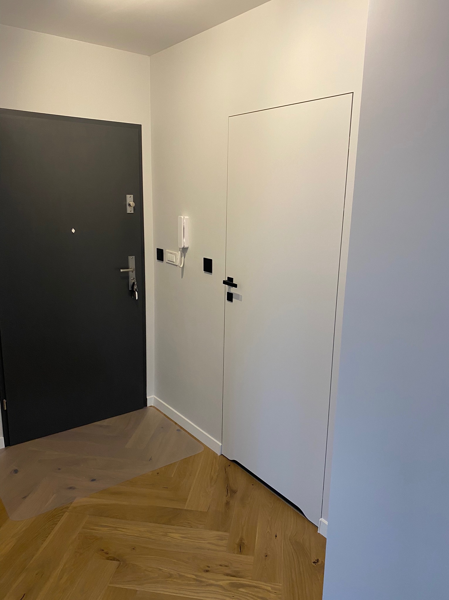 Korytarz po remoncie z dwiema drzwiami: czarne wejściowe z kluczami w zamku i białe wewnętrzne, parkiet ułożony w jodełkę, ściany pomalowane na biało.