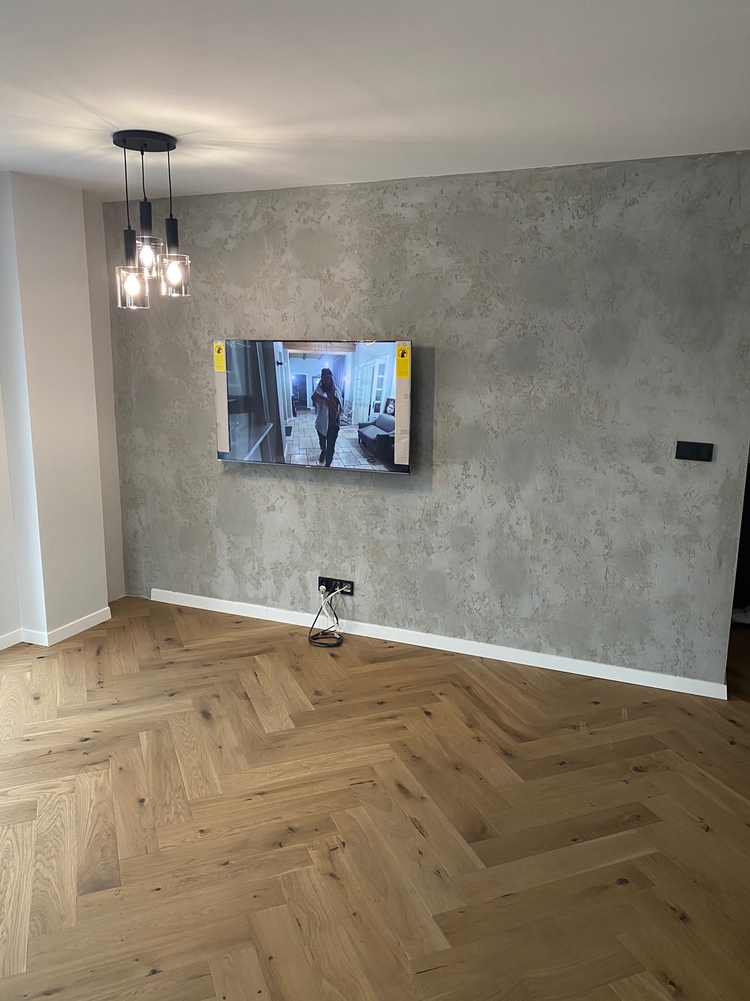 Salon z parkietem ułożonym w jodełkę, ściana z dekoracyjnym tynkiem, wiszący telewizor i nowoczesna lampa sufitowa z trzema szklanymi kloszami.