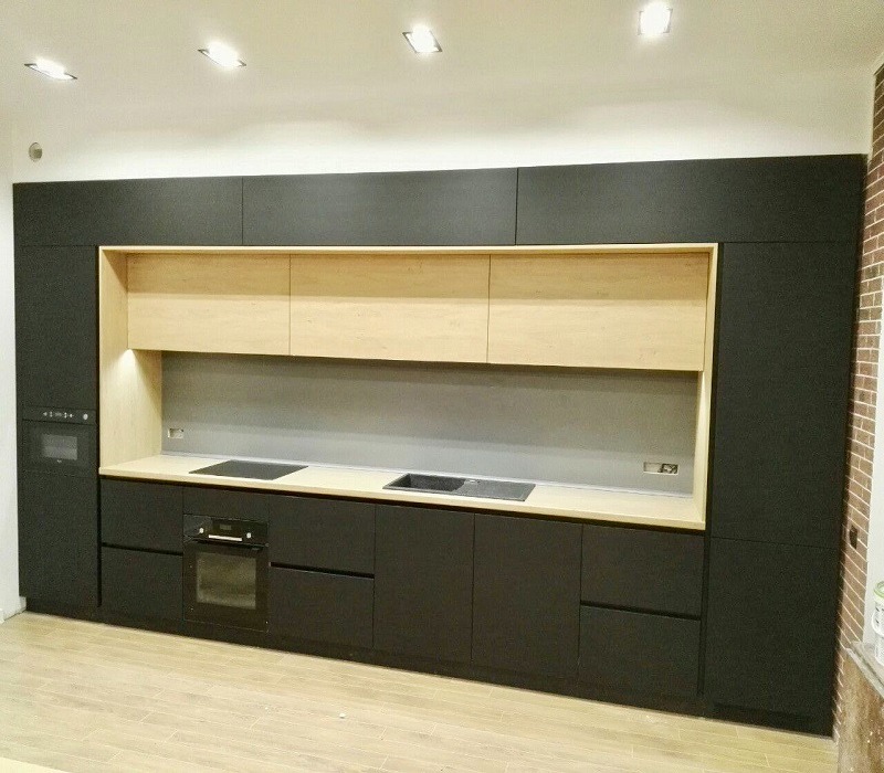 Nowoczesna kuchnia na wymiar z czarnymi frontami, jasnym drewnianym wykończeniem górnych szafek i blatem, płyta indukcyjna i piekarnik w zabudowie, widoczne oświetlenie sufitowe.