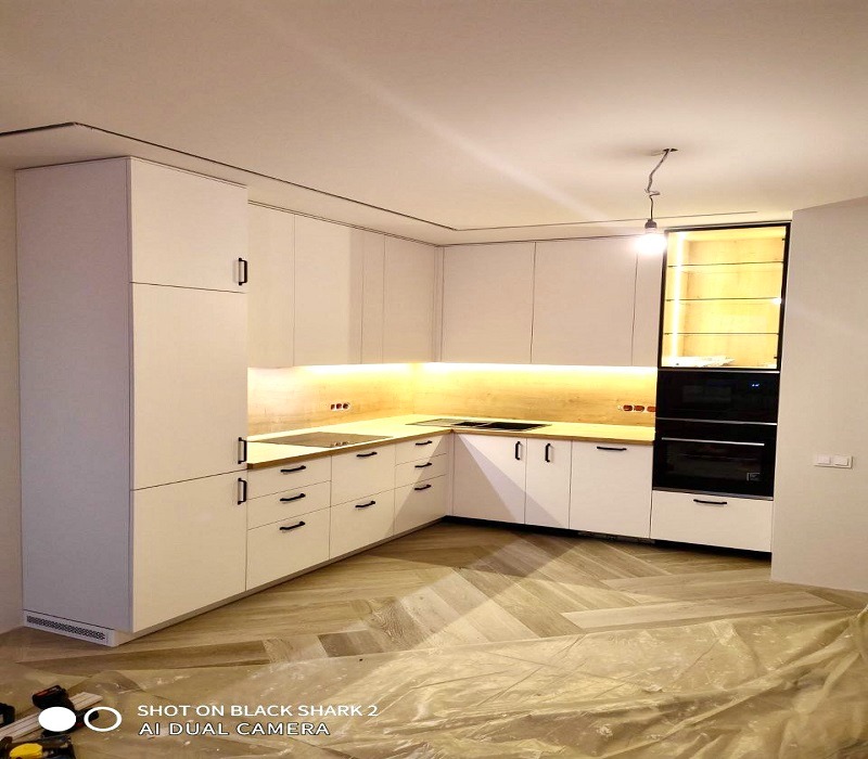 Jasna, narożna zabudowa kuchenna z drewnianym blatem i czarnymi uchwytami, oświetlenie LED pod szafkami wiszącymi, sprzęt AGD w zabudowie, podłoga ułożona w jodełkę, zabezpieczona folią malarską.