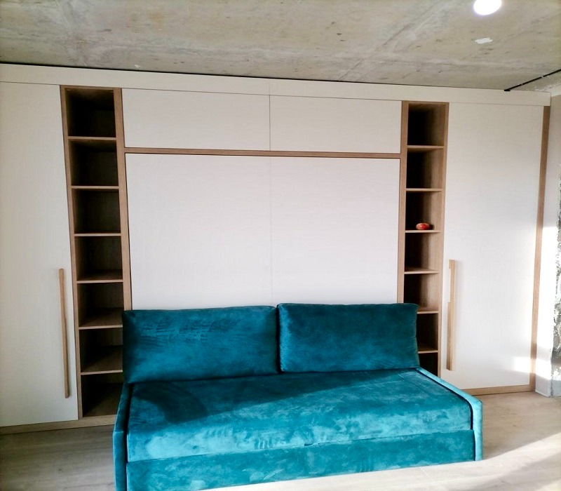 Biała szafa z rozkładanym łóżkiem i otwartymi półkami z drewnianymi akcentami oraz turkusową sofą na pierwszym planie, na tle surowego betonowego sufitu.