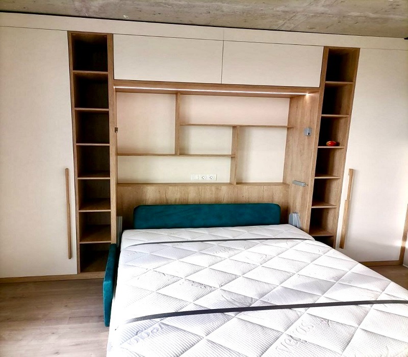 Złożone łóżko chowane w zabudowie meblowej z białymi szafami po bokach i drewnianą wnęką z półkami, oświetleniem LED i gniazdkami elektrycznymi nad wezgłowiem z turkusowym obiciem.