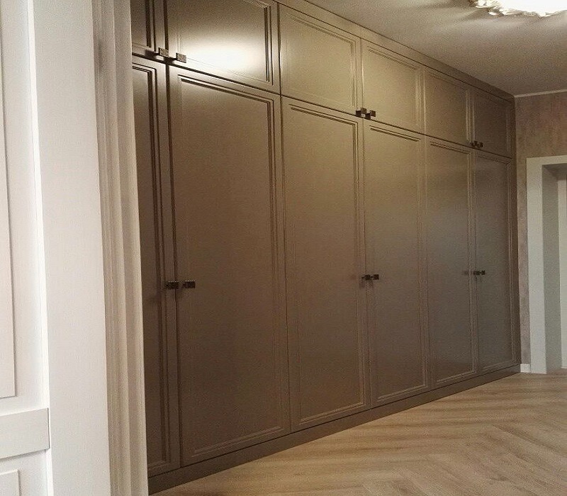 Wysoka, zabudowana szafa wnękowa z ciemnymi, kwadratowymi uchwytami i frezowanymi frontami, na podłodze parkiet ułożony w jodełkę.