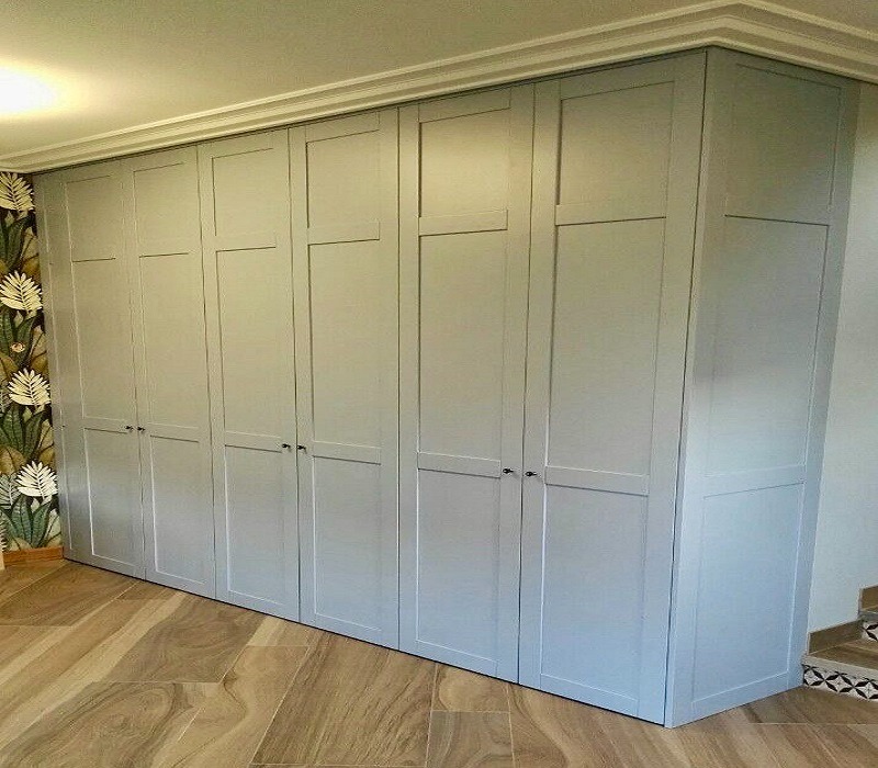 Szafa wnękowa na wymiar, szara, z siedmioma parami drzwi, z delikatnym frezowaniem, dopasowana do wysokości pomieszczenia, z dekoracyjną listwą przysufitową i tapetą w liście po lewej stronie.