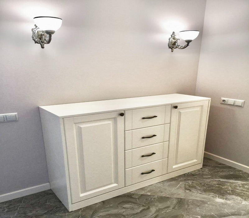 Biała komoda z czterema szufladami i dwojgiem drzwi, z ciemnymi uchwytami, oświetlona ozdobnymi kinkietami na ścianie, na podłodze z marmurowym wzorem.