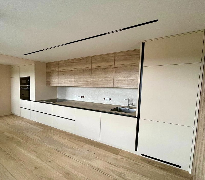 Nowoczesna kuchnia z białymi dolnymi szafkami i drewnopodobnymi górnymi, czarnym blatem i długim, czarnym oświetleniem sufitowym.