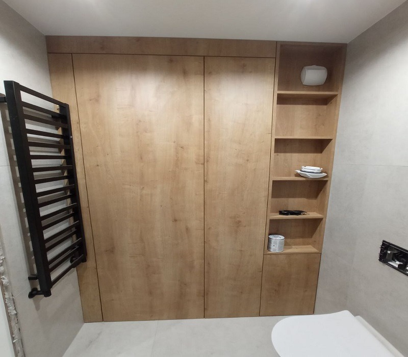 Drewniana zabudowa w łazience z szafą zamykaną na dwoje drzwi i otwartymi półkami, na których stoją akcesoria łazienkowe. Obok grzejnik drabinkowy.