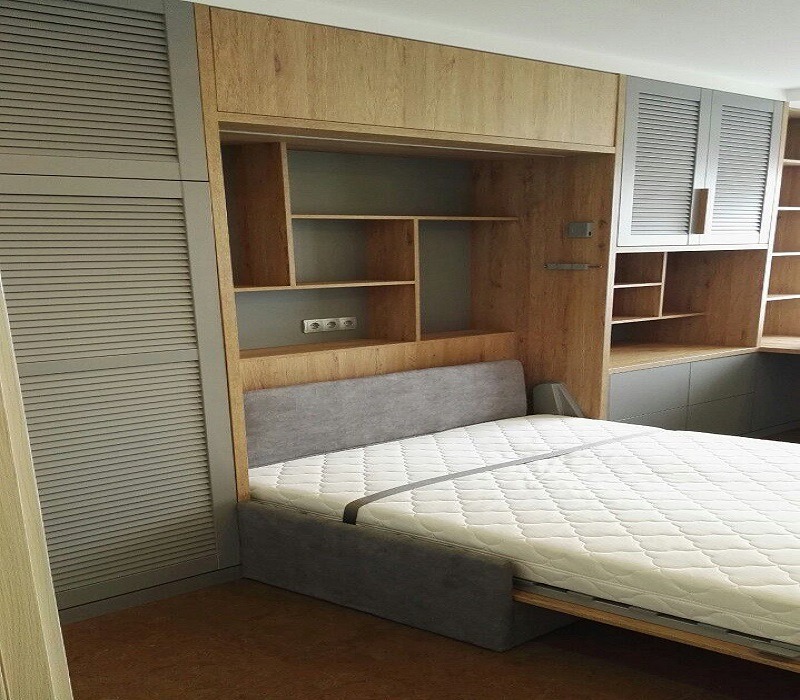 Szafa na wymiar z łóżkiem chowanym, jasne drewno i szare lamele, widoczne gniazdka elektryczne w zabudowie.