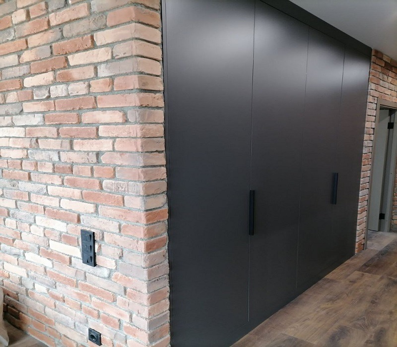 Czarna, matowa szafa wnękowa z minimalistycznymi uchwytami, wkomponowana w ścianę z cegły, z widocznymi gniazdkami elektrycznymi.