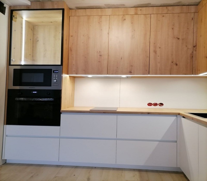 Nowoczesna zabudowa kuchenna z drewnianymi szafkami górnymi i białymi dolnymi, z wbudowanym piekarnikiem i mikrofalówką oraz oświetleniem LED podszafkowym, widoczne gniazdka elektryczne na ścianie.