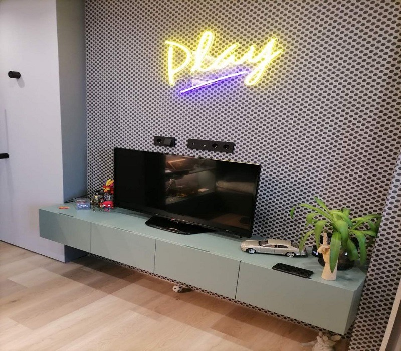 Nowoczesna szafka RTV w odcieniach pastelowej zieleni, zawieszona na ścianie z tapetą w drobne kropki, podświetlona neonem 'Play', z dekoracjami na blacie.
