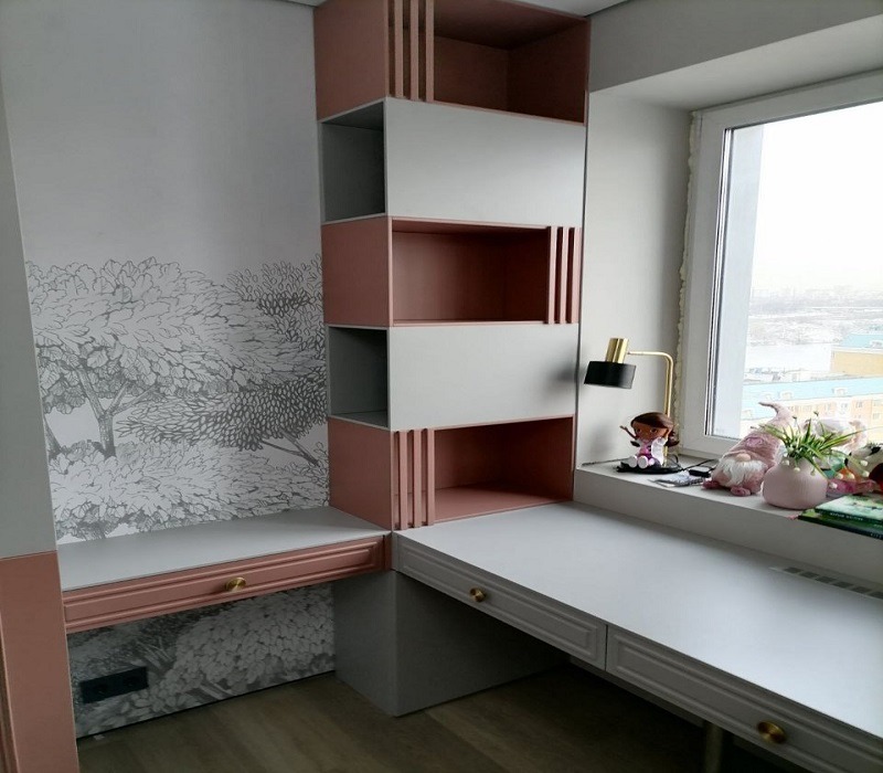 Biało-różowa zabudowa pokoju dziecięcego: biurko z szufladami i regał z otwartymi półkami, dopasowane do okna, z tapetą w leśny motyw w tle.