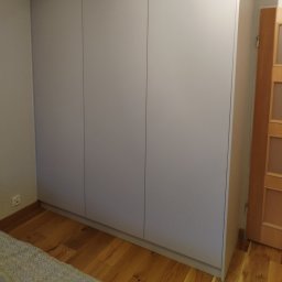 FUHP - Trzydrzwiowa szafa wnękowa z frontami w kolorze jasnoszarym, zlicowanymi z podłogą z drewnianych desek, widoczna w rogu pokoju obok drewnianych drzwi.