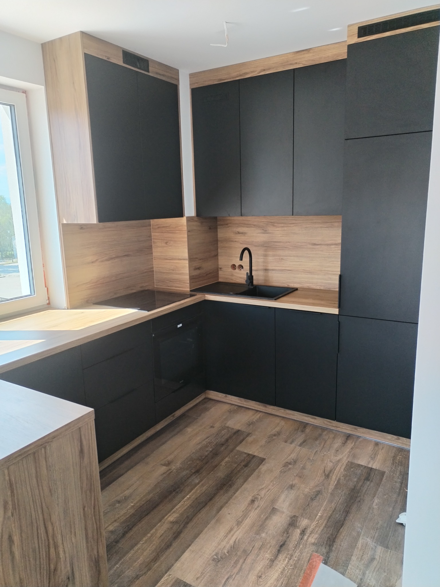 Nowoczesna kuchnia z czarnymi frontami i drewnopodobnymi blatami, widok na zabudowę z piekarnikiem i płytą grzewczą oraz zlewozmywakiem z czarną armaturą, drewniana podłoga.