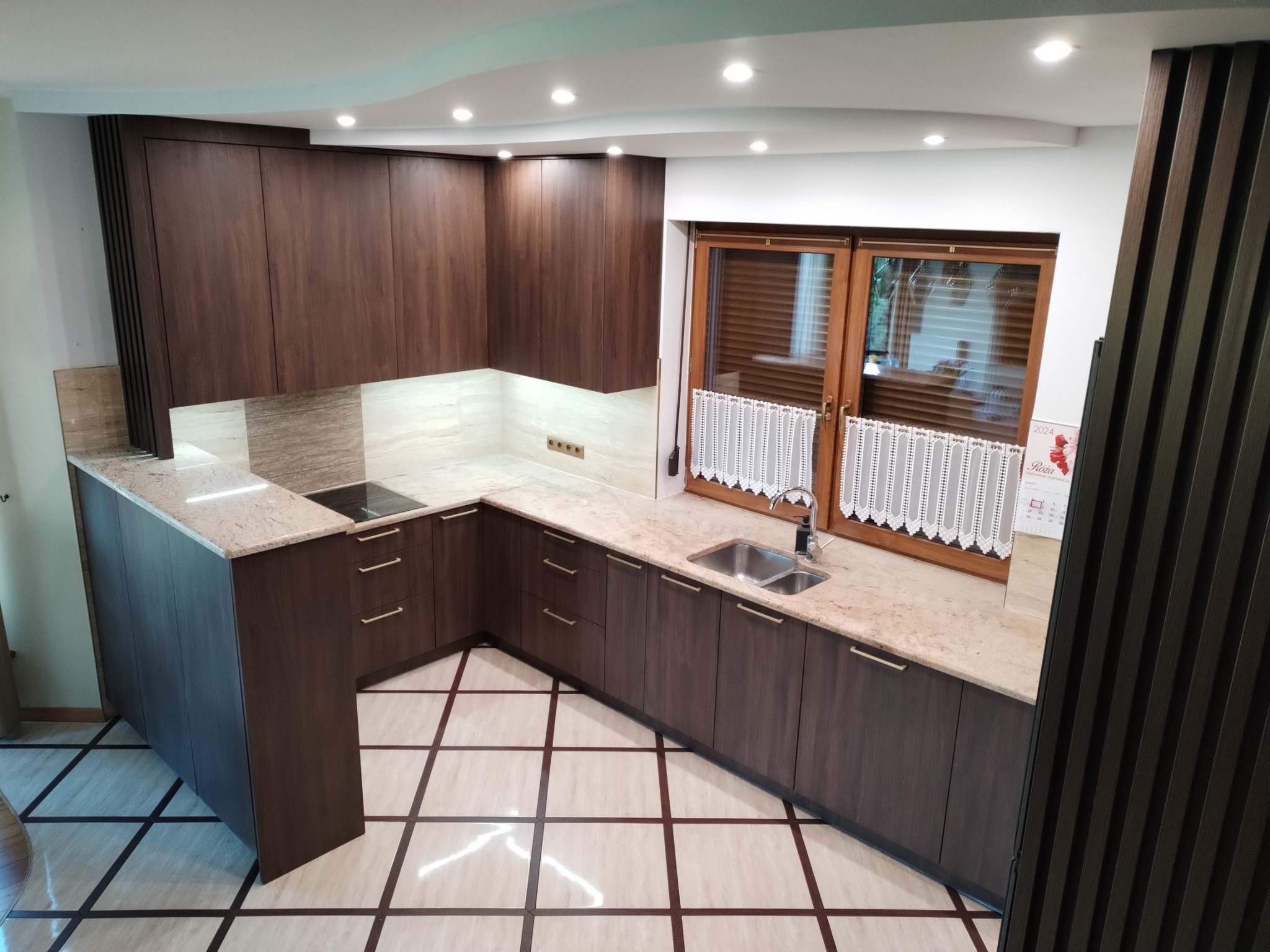 Nowoczesna kuchnia z ciemnobrązowymi szafkami, jasnym blatem i płytkami podłogowymi w kratkę. Okno z drewnianą ramą i białymi, koronkowymi firankami.