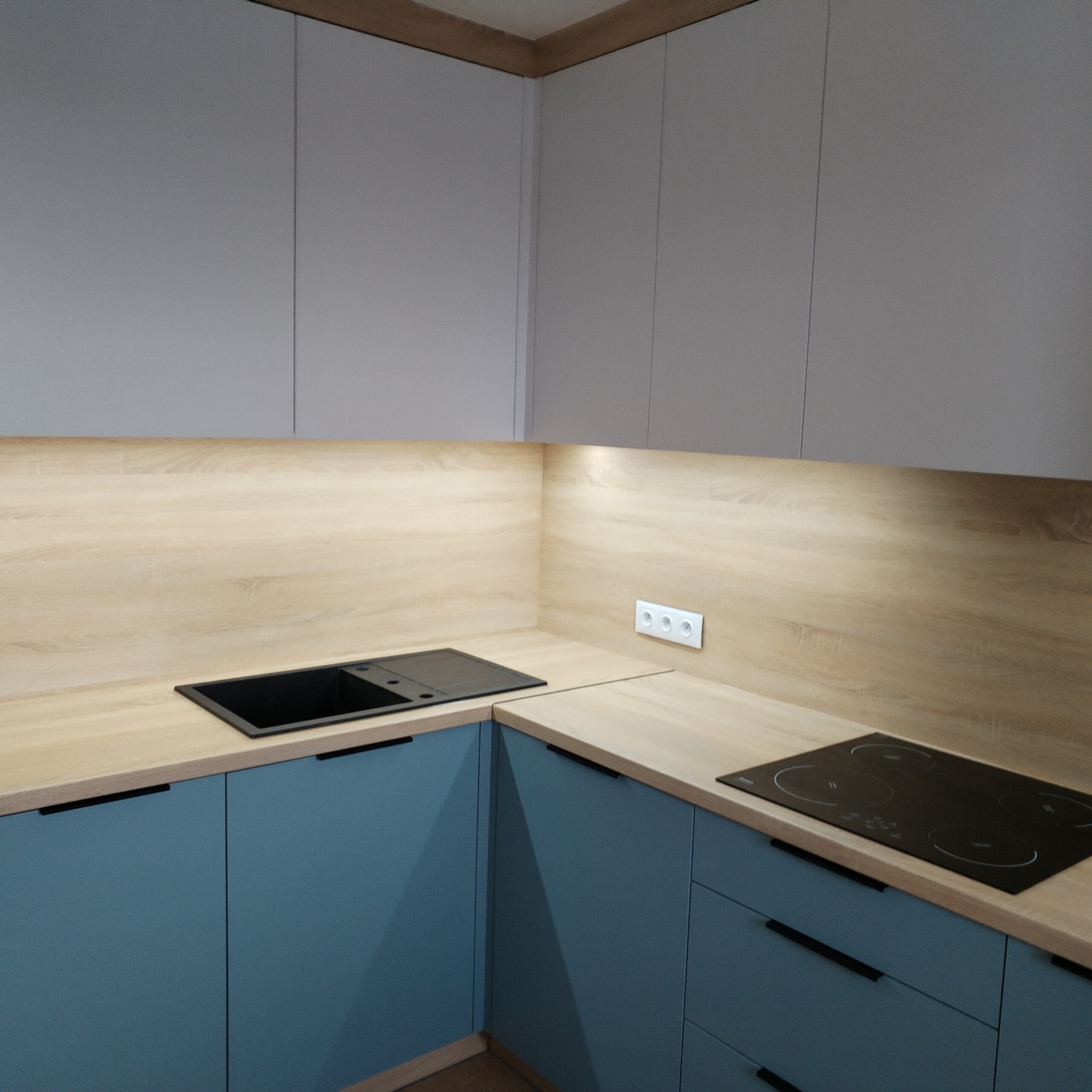 Nowoczesna kuchnia narożna z szarymi szafkami górnymi i niebieskimi dolnymi, blatem imitującym jasne drewno oraz czarnym zlewem i płytą indukcyjną.
