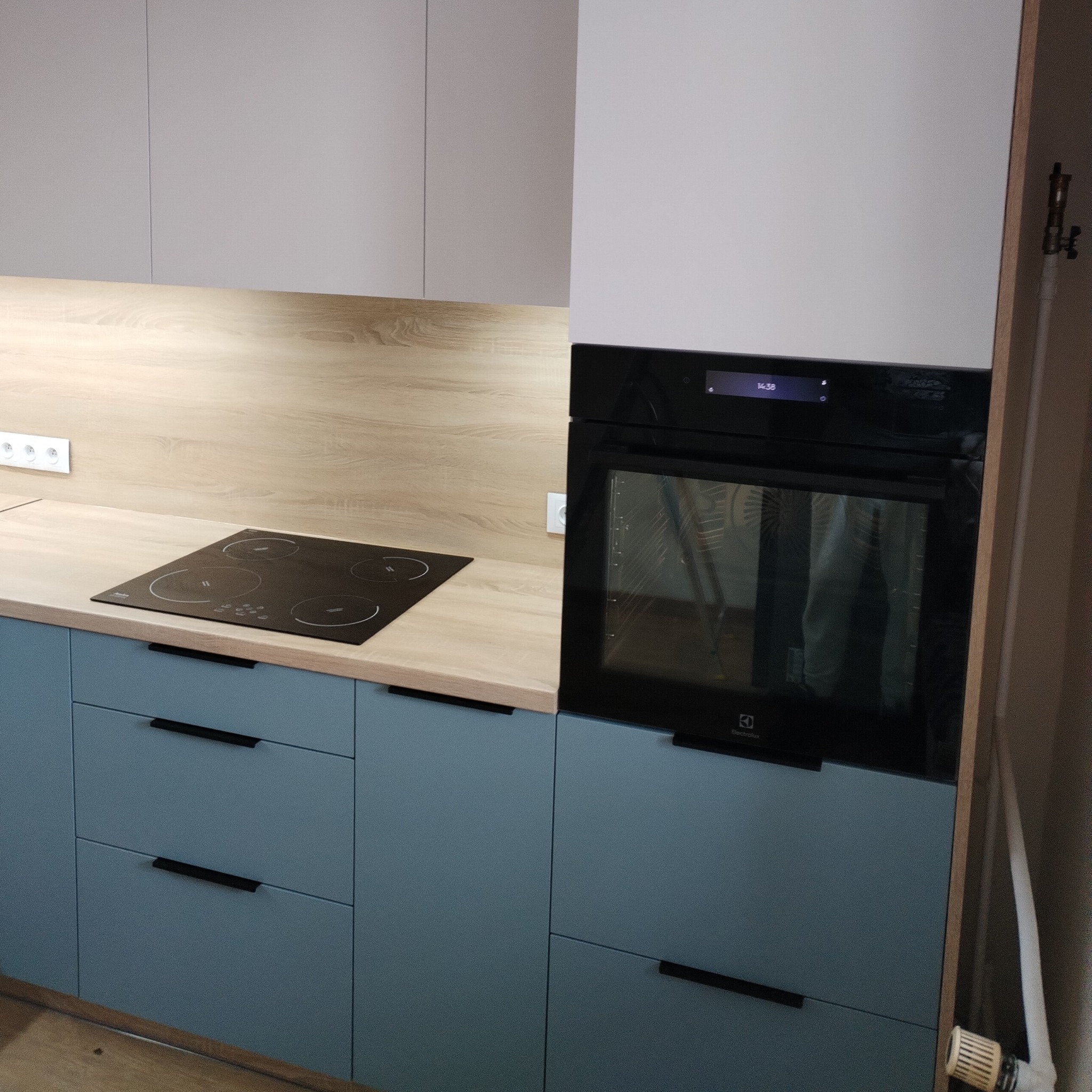 Fragment zabudowy kuchennej z niebieskimi frontami szafek, drewnianym blatem, czarną płytą indukcyjną i piekarnikiem Electrolux z widocznym wyświetlaczem.