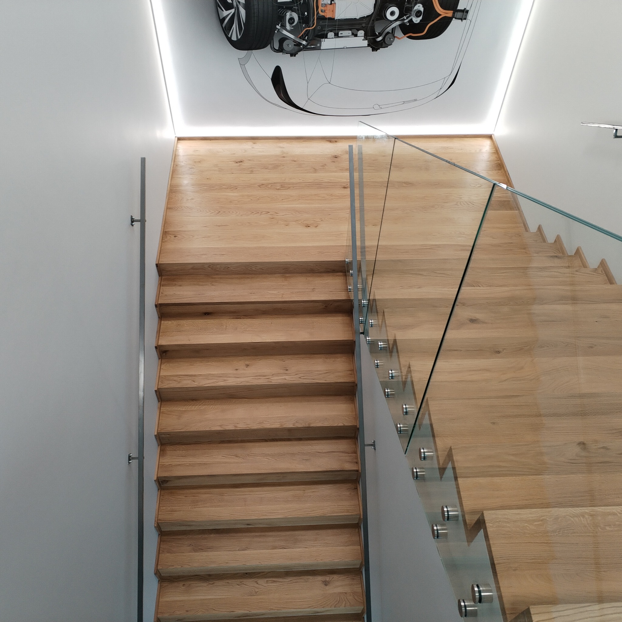 Jasne drewniane schody z szklaną balustradą, mocowaną metalowymi uchwytami; na suficie grafika przedstawiająca podwozie samochodu elektrycznego.