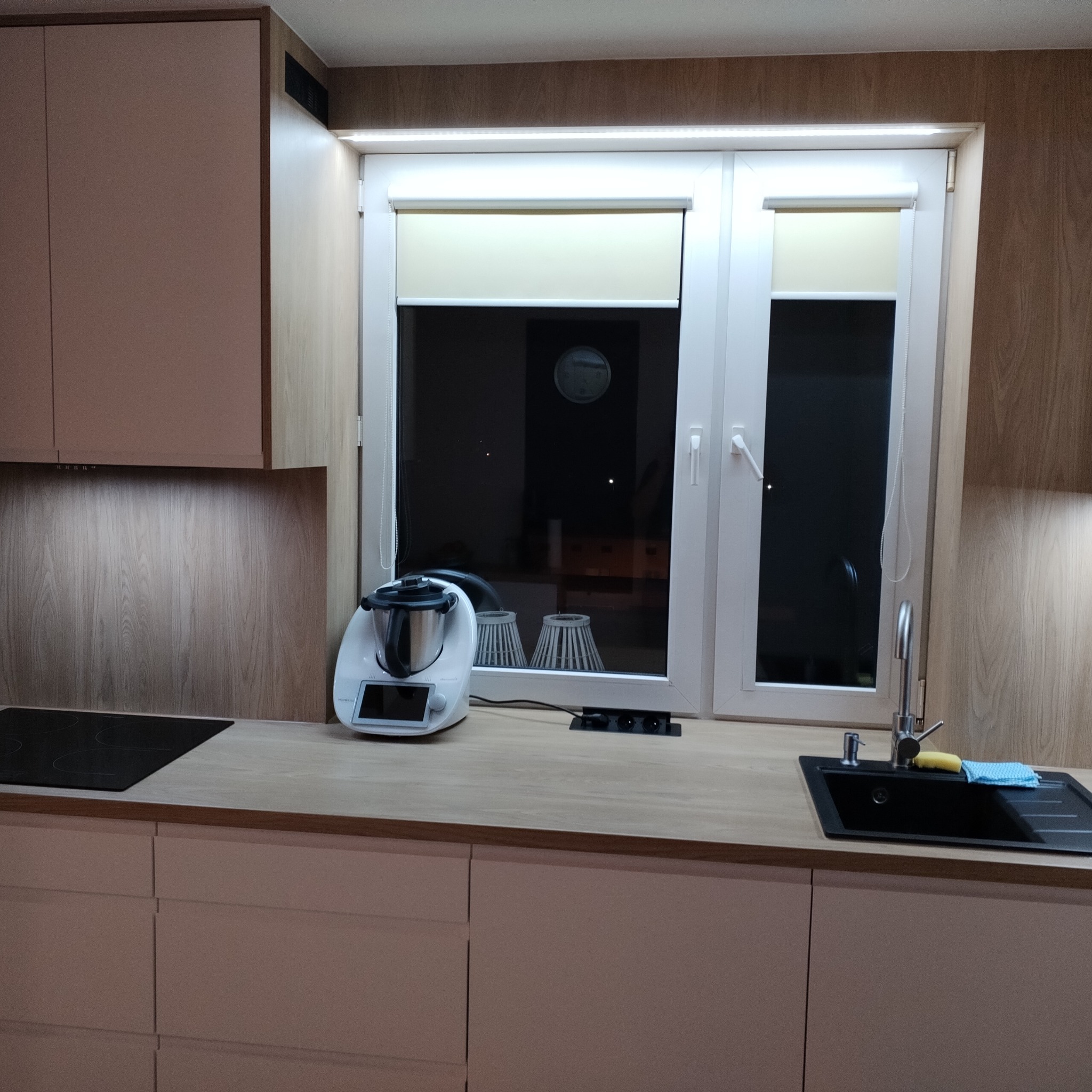 Nowoczesna kuchnia z beżowymi frontami, drewnopodobnym blatem i białym oknem z roletą, wyposażona w czarną płytę grzewczą, biały robot kuchenny i czarny zlew z baterią.