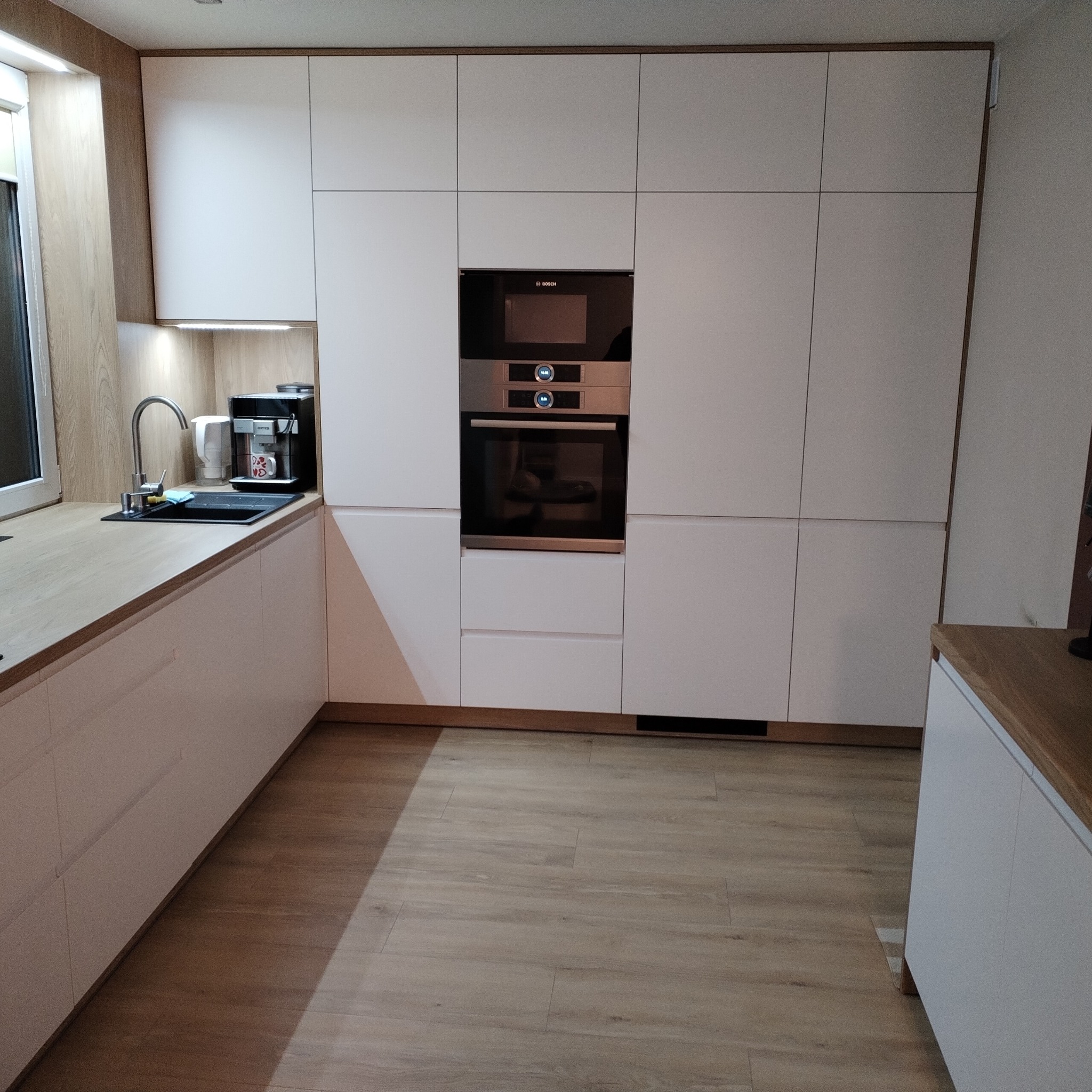 Nowoczesna kuchnia z białymi, gładkimi frontami szafek, drewnianym blatem i zabudowanym sprzętem AGD marki Bosch.