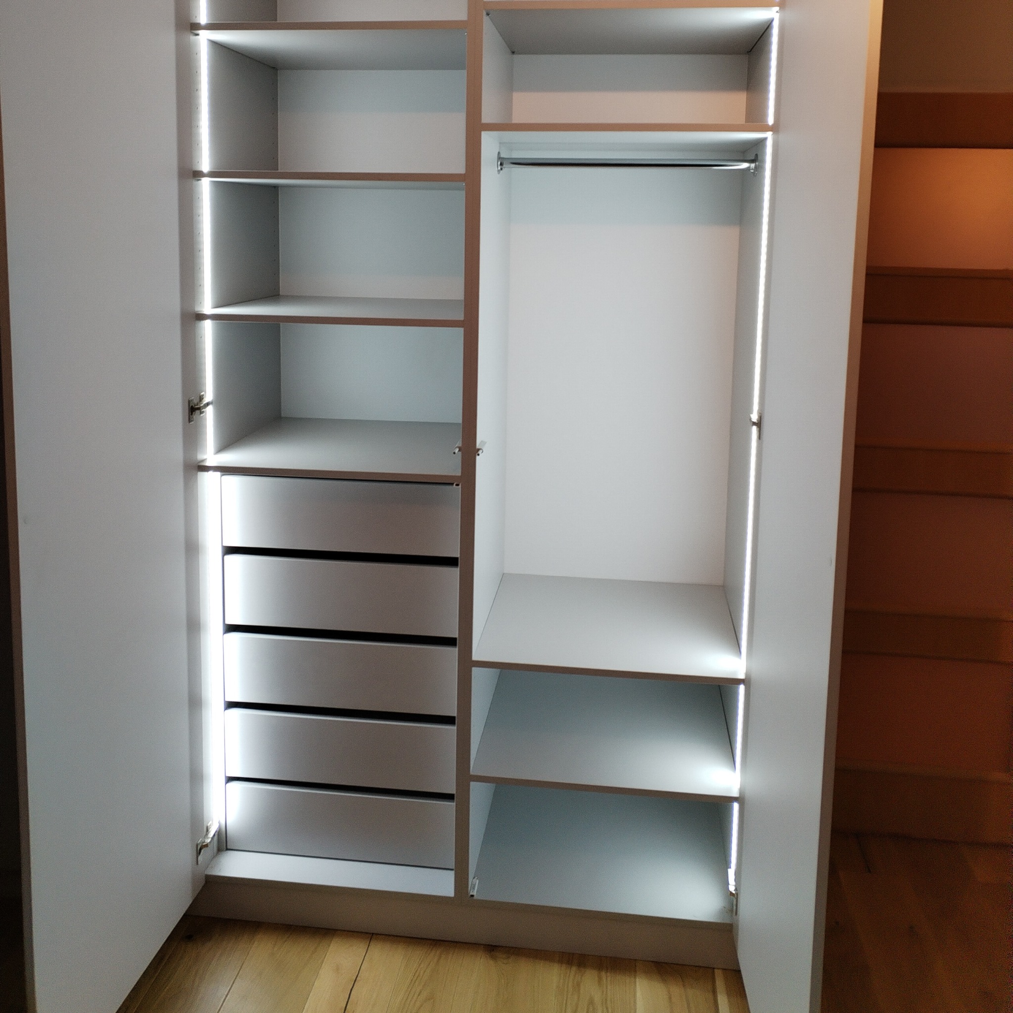 Otwarta, biała szafa z półkami, szufladami i drążkiem, oświetlona od wewnątrz ledowymi paskami, na drewnianej podłodze.