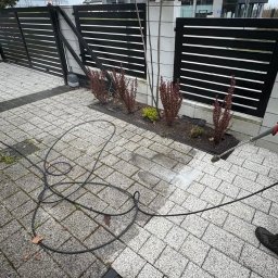SpecGreen - Czyszczenie kostki brukowej myjką ciśnieniową na posesji z czarnym ogrodzeniem panelowym i rabatami z roślinami ozdobnymi. Częściowo umyta powierzchnia.