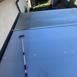 SpecGreen - Czyszczenie zadaszenia tarasu szczotką teleskopową. Widoczna różnica czystości na powierzchni. Tło: ogród i budynek.