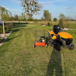 SpecGreen - Żółty traktorek do trawy i kosiarka na tle świeżo skoszonego trawnika w słoneczny dzień. Widoczne drzewa i krzewy w tle.